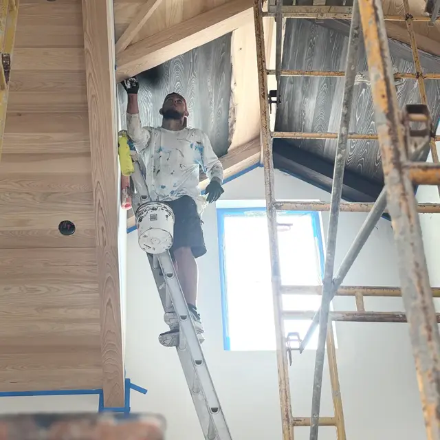 Painter on a ladder finishing a two-story interior wall below a wood ceiling